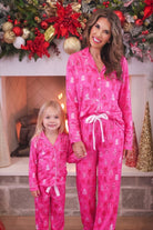 A woman and a young girl wearing matching bright pink Christmas-themed pajamas with white snowman and sleigh prints, standing in front of a decorated fireplace.