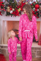 A woman and a young girl wearing matching bright pink Christmas-themed pajamas with a white bow tie, standing in front of a decorated fireplace.
