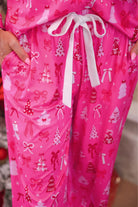 Bright pink pajama pants with a white drawstring waist and an all-over Christmas print featuring bows, gifts, and trees, shown on a person wearing matching top.