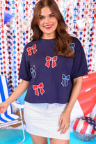 banded short-sleeved cropped fit luxe top with blue and red metallic foil bows outlined in white embroidery and side slits