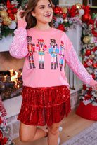 a lightweight pink long-sleeve top decorated with four vibrant nutcracker designs finished with dimensional beaded effect boots and accented by soft, shimmering velvet-sequin sleeves, a relaxed drop shoulder, and ribbed crew neckline, cuffs, and hem