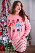 a lightweight pink long-sleeve top decorated with four vibrant nutcracker designs finished with dimensional beaded effect boots and accented by soft, shimmering velvet-sequin sleeves, a relaxed drop shoulder, and ribbed crew neckline, cuffs, and hem