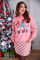 a lightweight pink long-sleeve top decorated with four vibrant nutcracker designs finished with dimensional beaded effect boots and accented by soft, shimmering velvet-sequin sleeves, a relaxed drop shoulder, and ribbed crew neckline, cuffs, and hem
