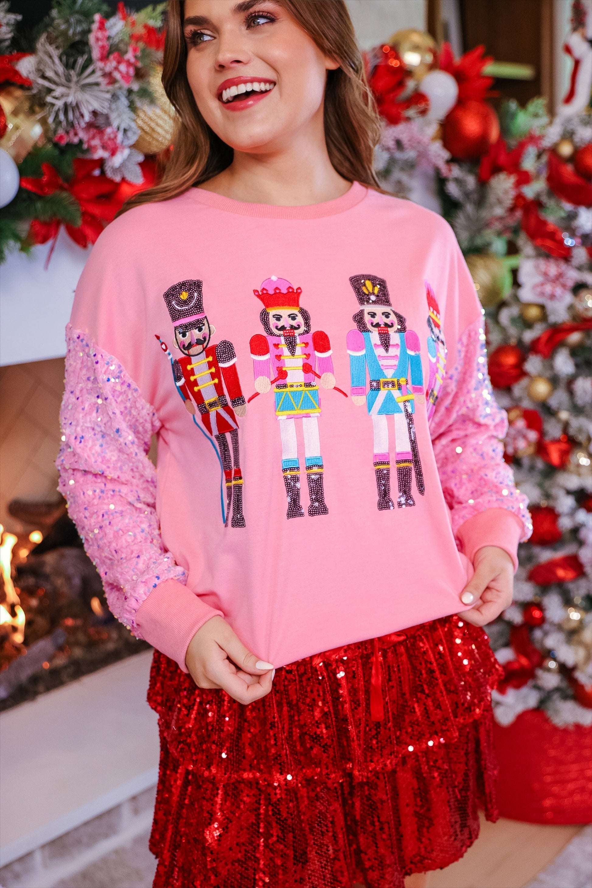 a lightweight pink long-sleeve top decorated with four vibrant nutcracker designs finished with dimensional beaded effect boots and accented by soft, shimmering velvet-sequin sleeves, a relaxed drop shoulder, and ribbed crew neckline, cuffs, and hem