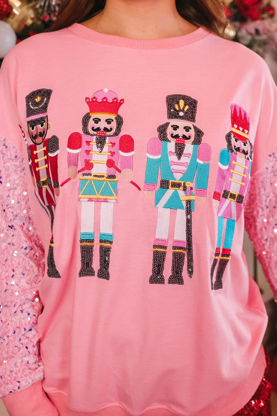 a lightweight pink long-sleeve top decorated with four vibrant nutcracker designs finished with dimensional beaded effect boots and accented by soft, shimmering velvet-sequin sleeves, a relaxed drop shoulder, and ribbed crew neckline, cuffs, and hem