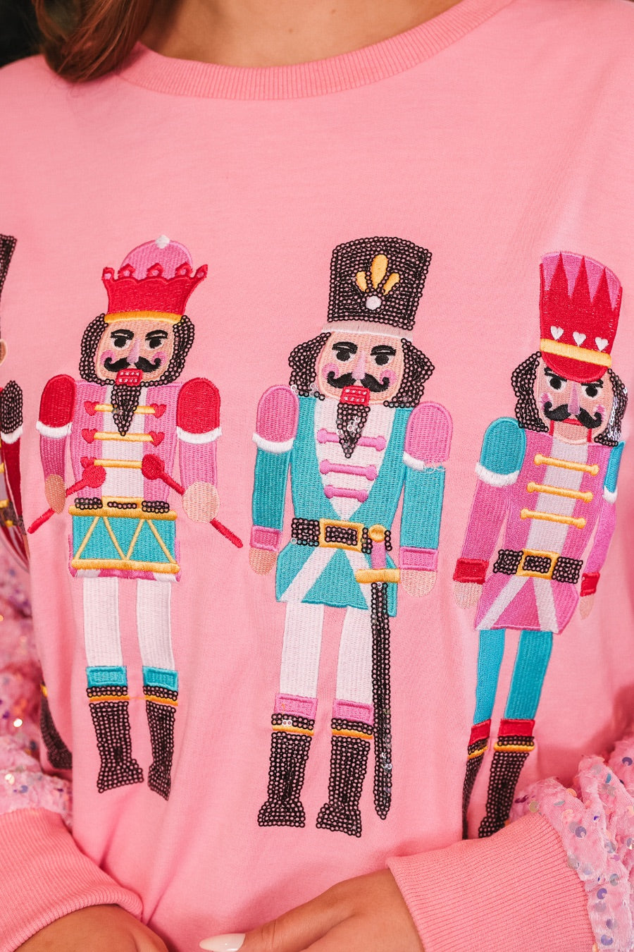 a lightweight pink long-sleeve top decorated with four vibrant nutcracker designs finished with dimensional beaded effect boots and accented by soft, shimmering velvet-sequin sleeves, a relaxed drop shoulder, and ribbed crew neckline, cuffs, and hem