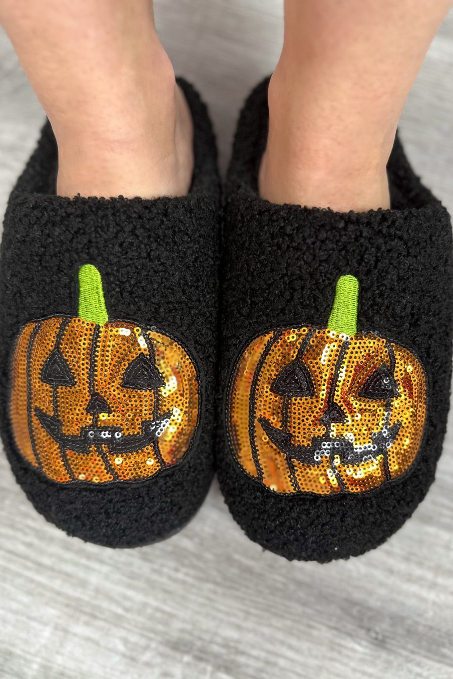 black fuzzy mule slippers with orange sequin jack-o'-lantern appliqués, embroidered green stem accents, cushioned flatbeds, plush lining, open back design, and textured bottoms
