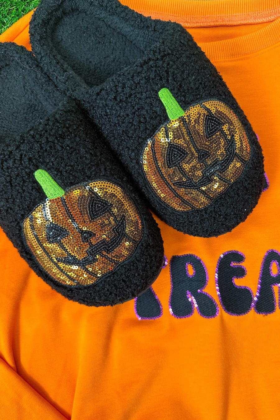 black fuzzy mule slippers with orange sequin jack-o'-lantern appliqués, embroidered green stem accents, cushioned flatbeds, plush lining, open back design, and textured bottoms