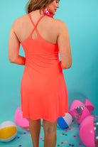 a coral sleeveless tennis dress designed with built-in shorts featuring hidden side pockets, adjustable crisscross straps, and built-in shelf bra