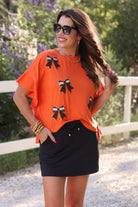 fluttery ruffle short-sleeve top with a mom crop length, sewn-on chenille football bow patches outlined in sequins on a soft, stretch-knit fabric in orange
