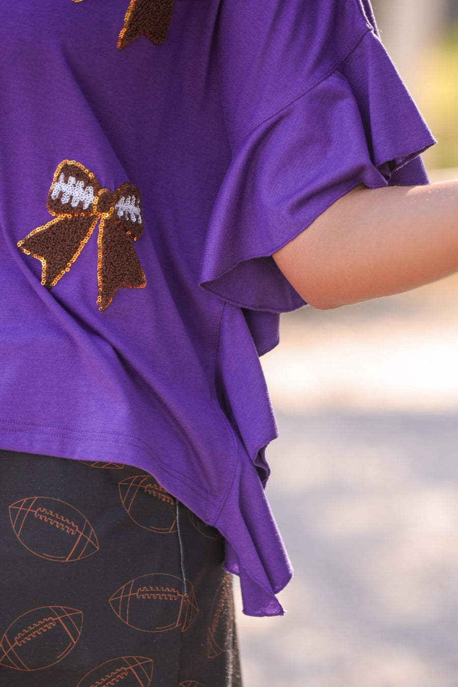 fluttery ruffle short-sleeve top with a mom crop length, sewn-on chenille football bow patches outlined in sequins on a soft, stretch-knit fabric in purple