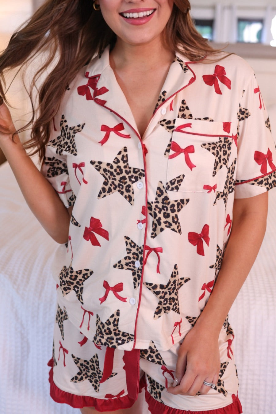 short-sleeve button-down style top with contrast piping and a chest pocket, paired with matching ruffle-trimmed shorts with an elastic waist and decorative tie, designed on a leopard star and red bow pattern print and a cream base, crafted on a soft, lightweight stretchy fabric