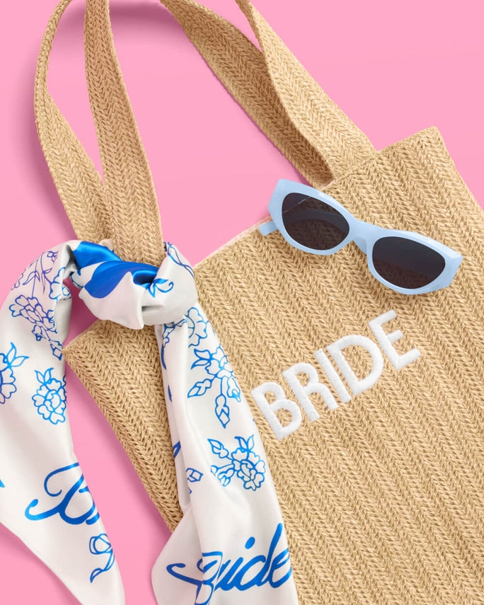 Beach bag with 'BRIDE' text, sunglasses, and floral scarf on a pink background