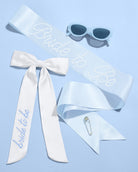 White sash with 'bride to be' text, sunglasses, and a safety pin on a light blue background