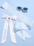 White sash with 'bride to be' text, sunglasses, and a brooch on a light blue background