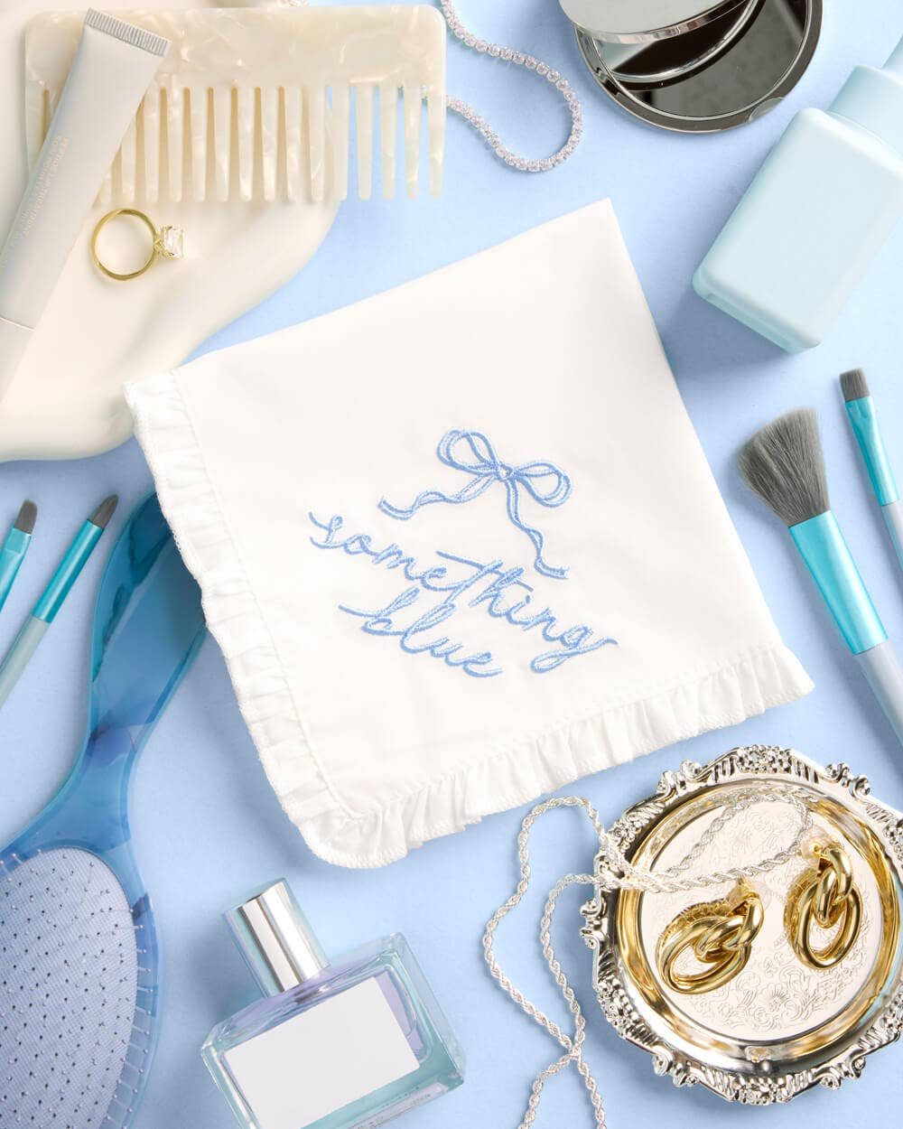 Collection of bridal accessories including a necklace, earrings, perfume, and a 'Something Blue' bag on a light blue background.