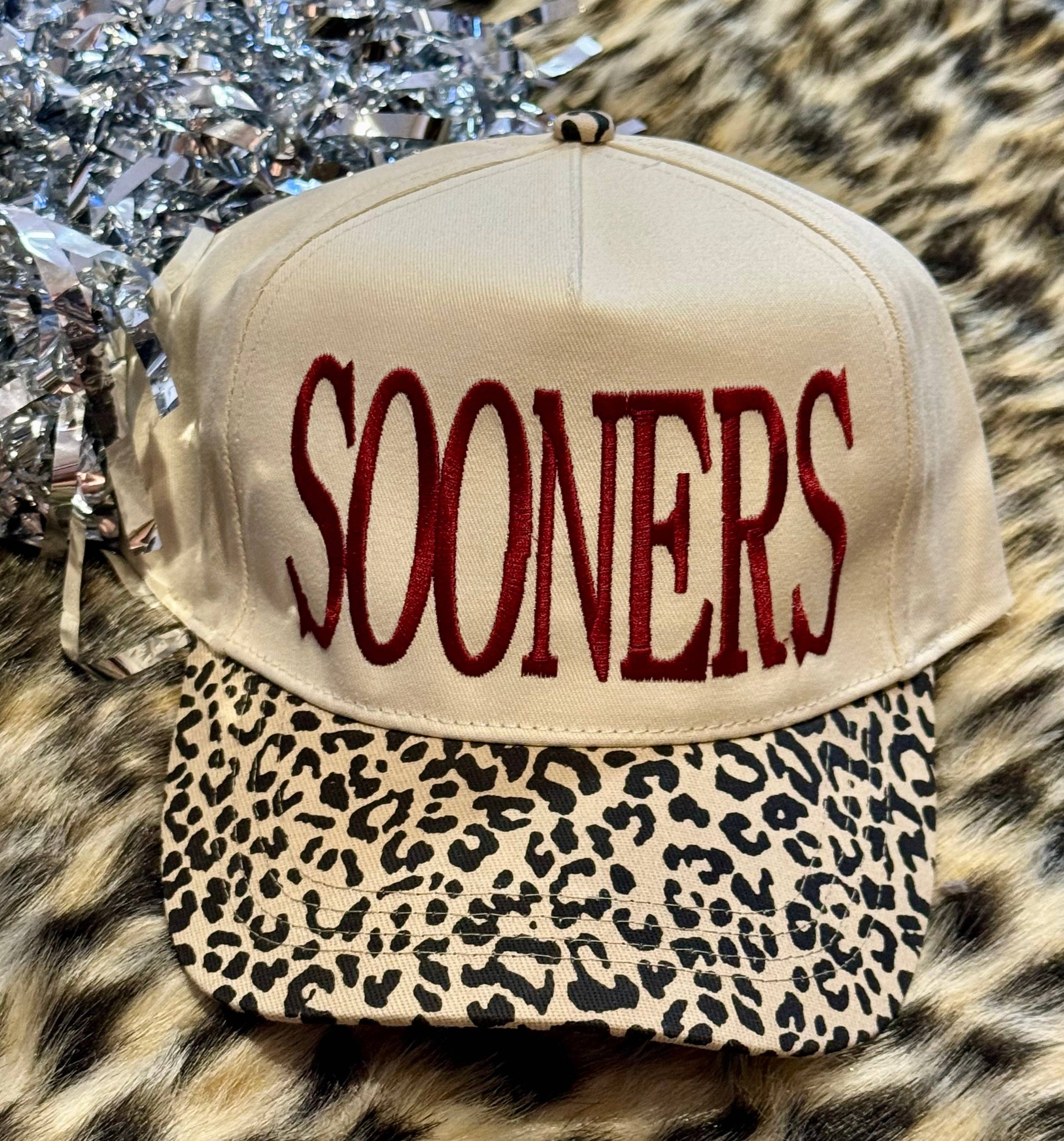 a lightweight unisex two-tone hat with embroidered crimson 'SOONERS' lettering, cream structured front, curved contrasting black leopard bill, coordinating leopard top button, and adjustable snap-back closure