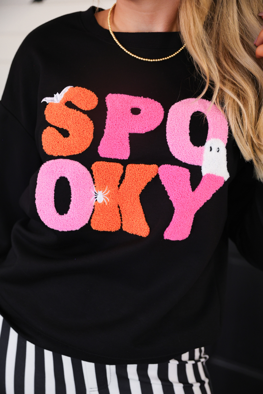 a black long-sleeve pullover made of ultra soft luxe material with bold plush chenille 'SPOOKY' patch lettering (orange and pink), white embroidered character accents (tiny bat, ghost and spider), ribbed cuffs, a banded hem, and a relaxed fit for a classic silhouette
