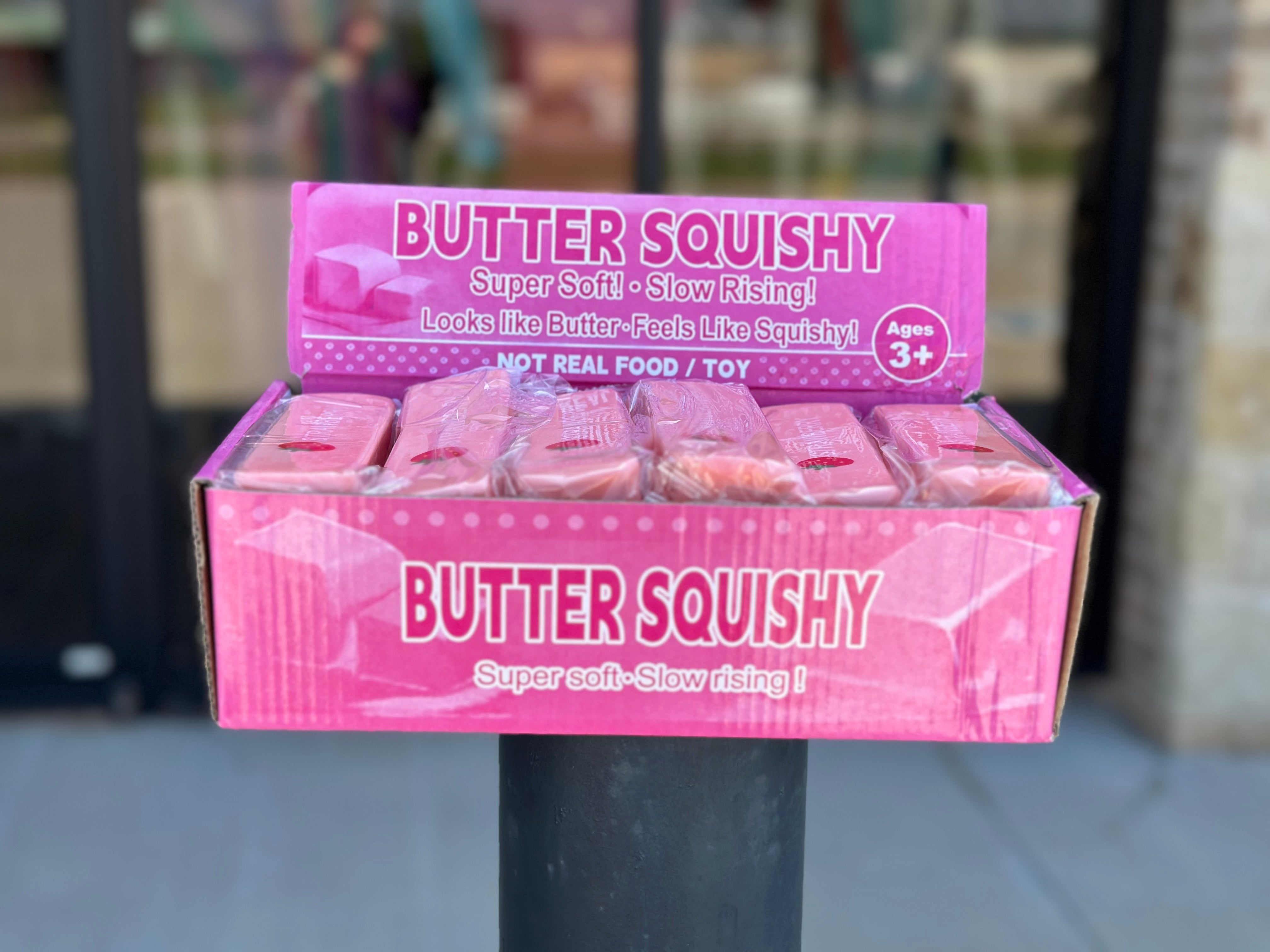 A display box of Butter Squishy toys on a black post outdoors. The box is pink with white text reading 'BUTTER SQUISHY' and shows several individually wrapped pink squishy toys inside.