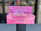 A display box of Butter Squishy toys on a black post outdoors. The box is pink with white text reading 'BUTTER SQUISHY' and shows several individually wrapped pink squishy toys inside.