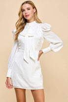 a white lined studded sheath dress with a high neckline accented with a decorative bow, puff shoulders, long cuffed sleeves, and a hidden rear zipper