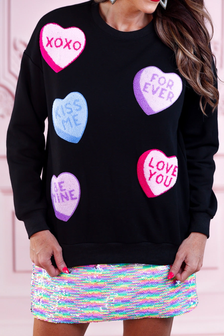 a black long-sleeve sweater made of ultra-soft material adorned with multicolor chenille patch conversation hearts, a relaxed drop-shoulder silhouette, and ribbed trim at the neckline, cuffs, and hemline