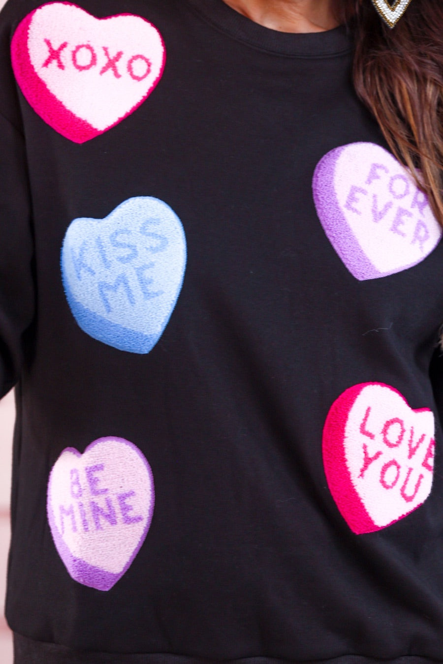 a black long-sleeve sweater made of ultra-soft material adorned with multicolor chenille patch conversation hearts, a relaxed drop-shoulder silhouette, and ribbed trim at the neckline, cuffs, and hemline