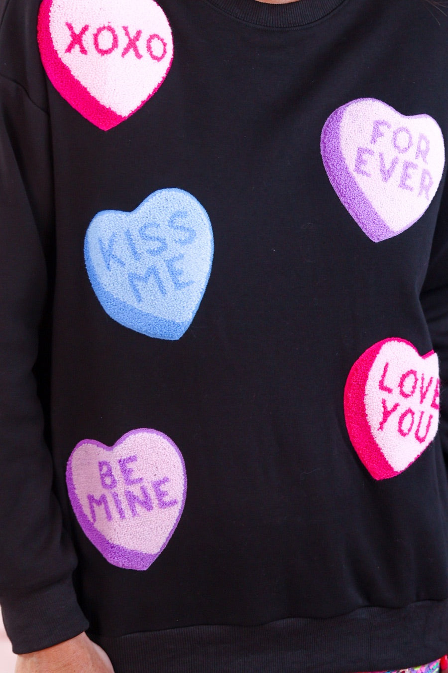 a black long-sleeve sweater made of ultra-soft material adorned with multicolor chenille patch conversation hearts, a relaxed drop-shoulder silhouette, and ribbed trim at the neckline, cuffs, and hemline