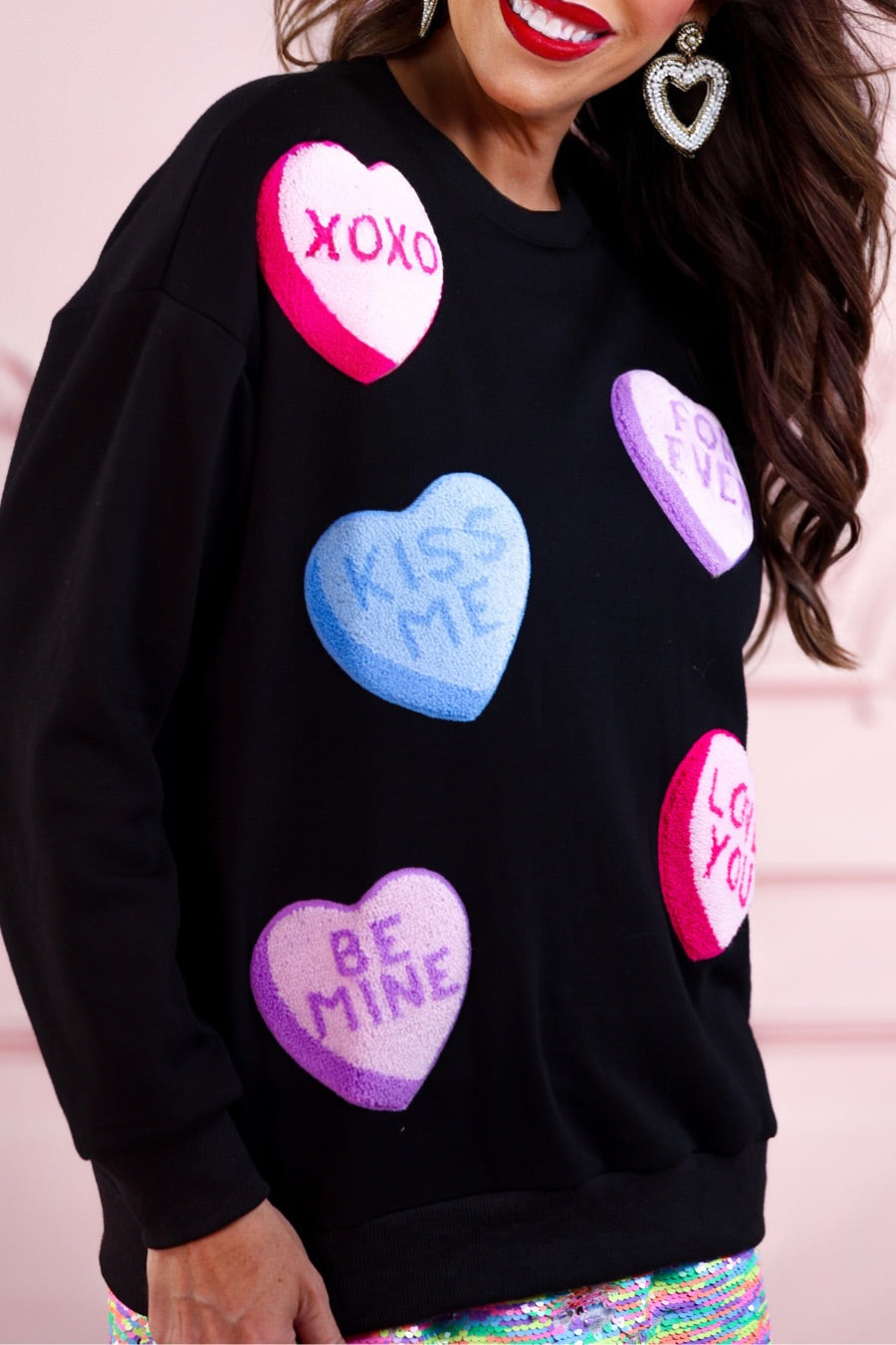 a black long-sleeve sweater made of ultra-soft material adorned with multicolor chenille patch conversation hearts, a relaxed drop-shoulder silhouette, and ribbed trim at the neckline, cuffs, and hemline