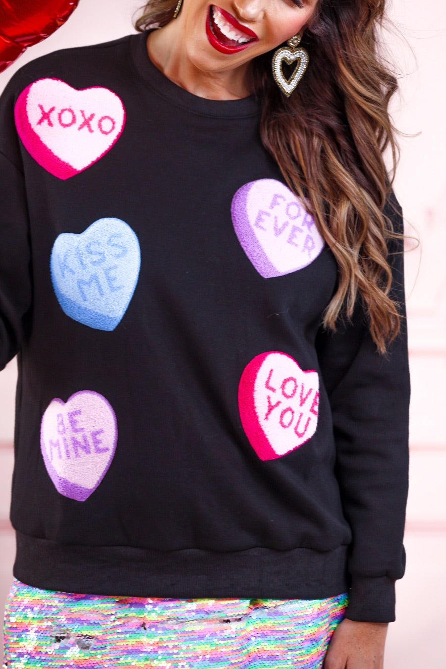 a black long-sleeve sweater made of ultra-soft material adorned with multicolor chenille patch conversation hearts, a relaxed drop-shoulder silhouette, and ribbed trim at the neckline, cuffs, and hemline