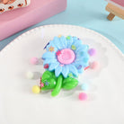 a blue flower-shaped decompression squishy toy with glossy petals, green leaves, mini decorative accents, and pastel pom poms