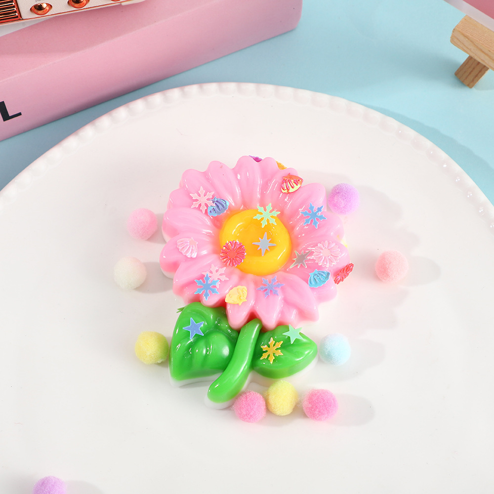 a pink flower-shaped decompression squishy toy with glossy petals, green leaves, mini decorative accents, and pastel pom poms