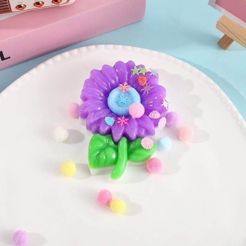 a purple flower-shaped decompression squishy toy with glossy petals, green leaves, mini decorative accents, and pastel pom poms