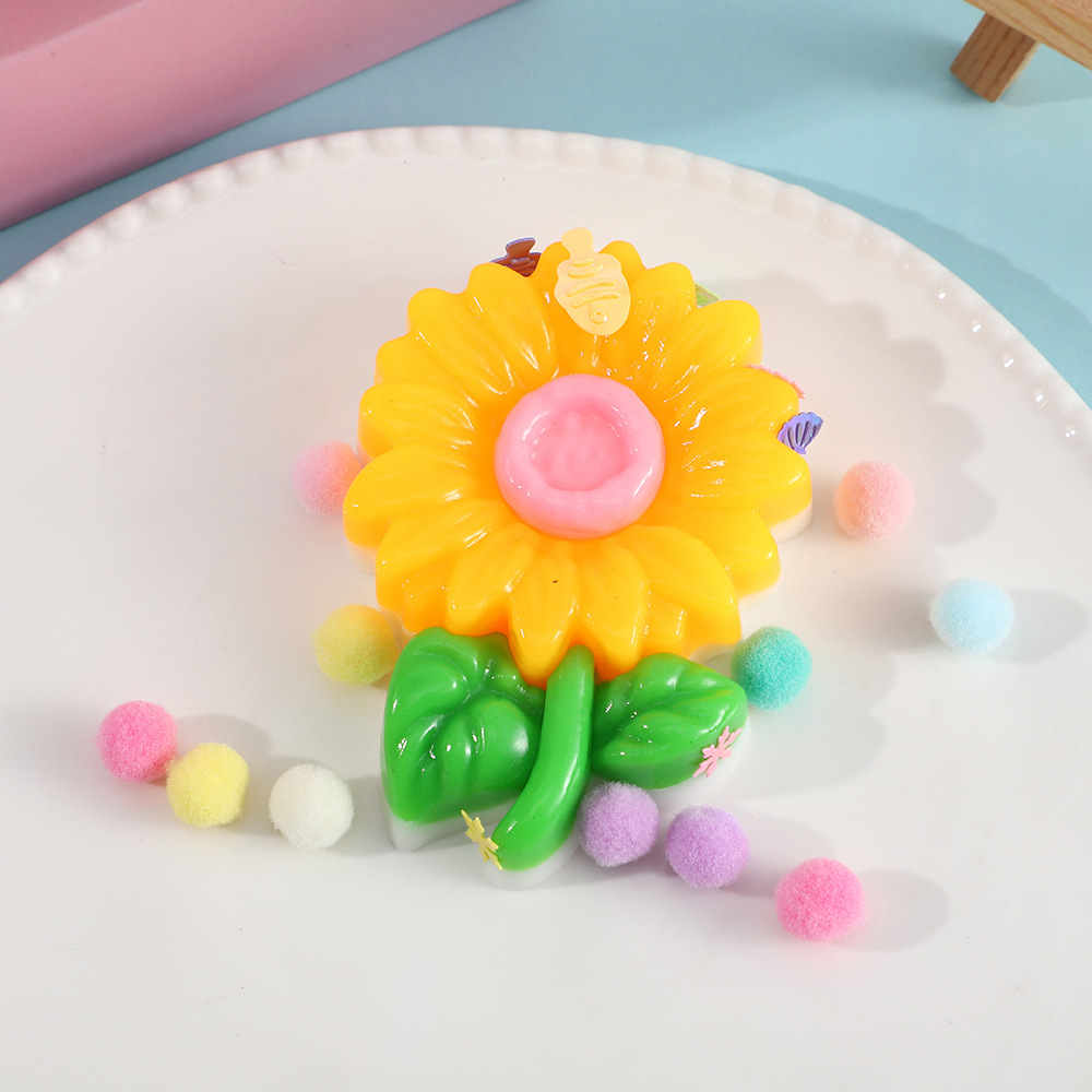 a yellow flower-shaped decompression squishy toy with glossy petals, green leaves, mini decorative accents, and pastel pom poms