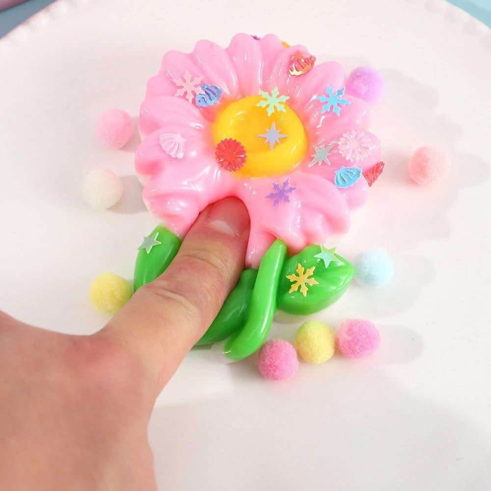a pink flower-shaped decompression squishy toy with glossy petals, green leaves, mini decorative accents, and pastel pom poms