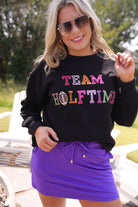 black long-sleeve sweatshirt made from soft mid-weight fabric with a crew neckline, straight hem, and bold multicolored varsity-style chenille patches that spell out 'TEAM HALFTIME' across the front chest