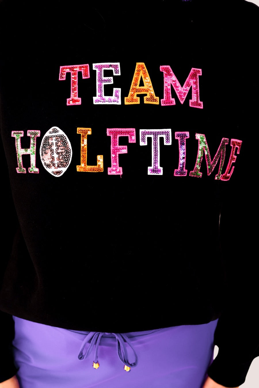 black long-sleeve sweatshirt made from soft mid-weight fabric with a crew neckline, straight hem, and bold multicolored varsity-style chenille patches that spell out 'TEAM HALFTIME' across the front chest