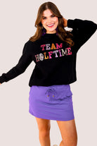 black long-sleeve sweatshirt made from soft mid-weight fabric with a crew neckline, straight hem, and bold multicolored varsity-style chenille patches that spell out 'TEAM HALFTIME' across the front chest