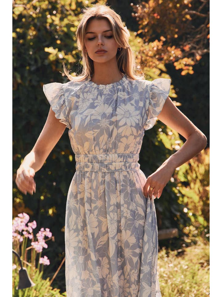 Woman wearing a floral dress standing in a garden with greenery and flowers.
