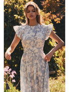 Woman wearing a floral dress standing in a garden with greenery and flowers.