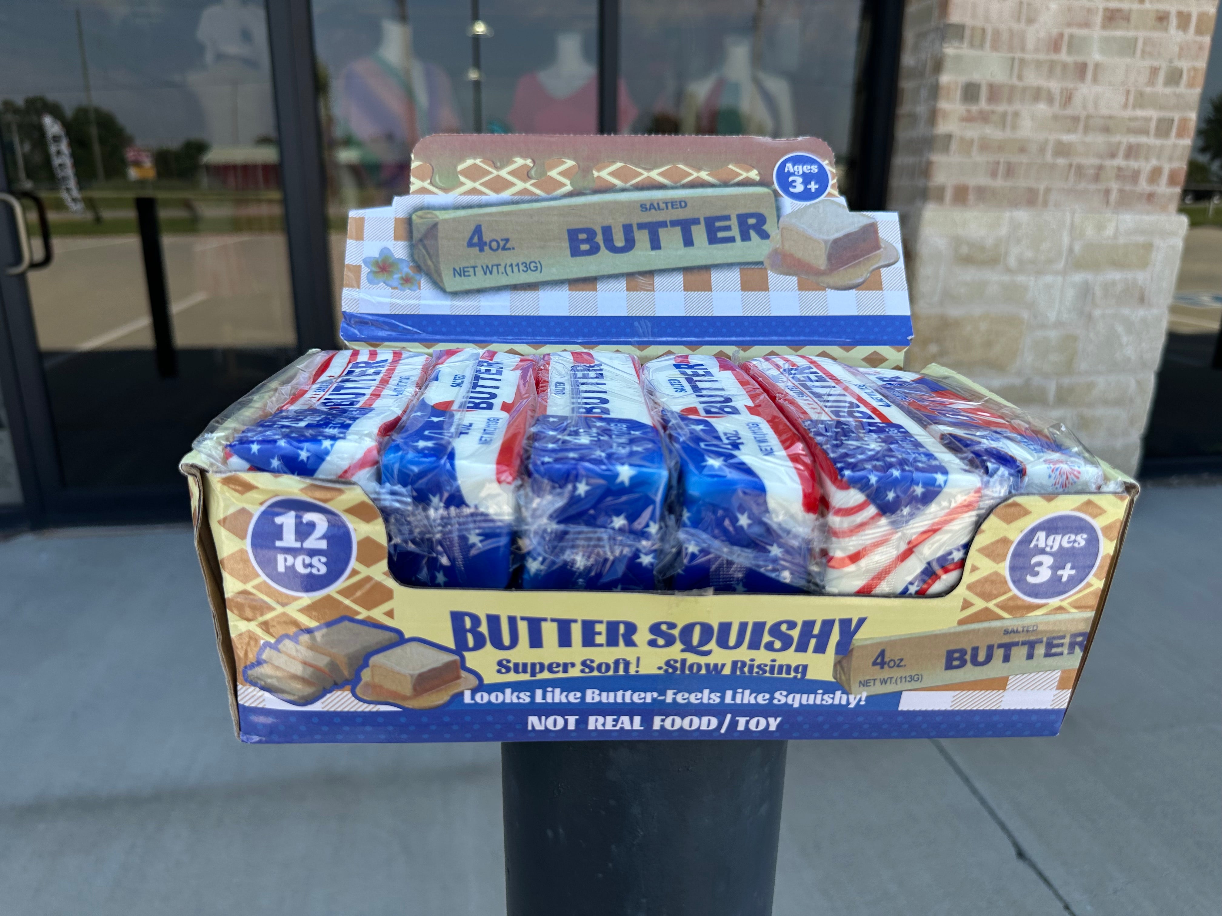 A retail display box filled with individually wrapped Butter Squishy toys, with a smaller display box placed behind it. The packaging features an American flag design with red, white, and blue, and text reading 'BUTTER SQUISHY' and '12 PCS'.