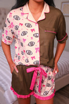 a short-sleeve button-down pajama top with a cream football and bow print, brown colorblock accents, pink contrast piping, and a chest pocket, paired with ruffle trimmed shorts, an elastic waistband, and a decorative tie, crafted on soft lightweight stretch fabric