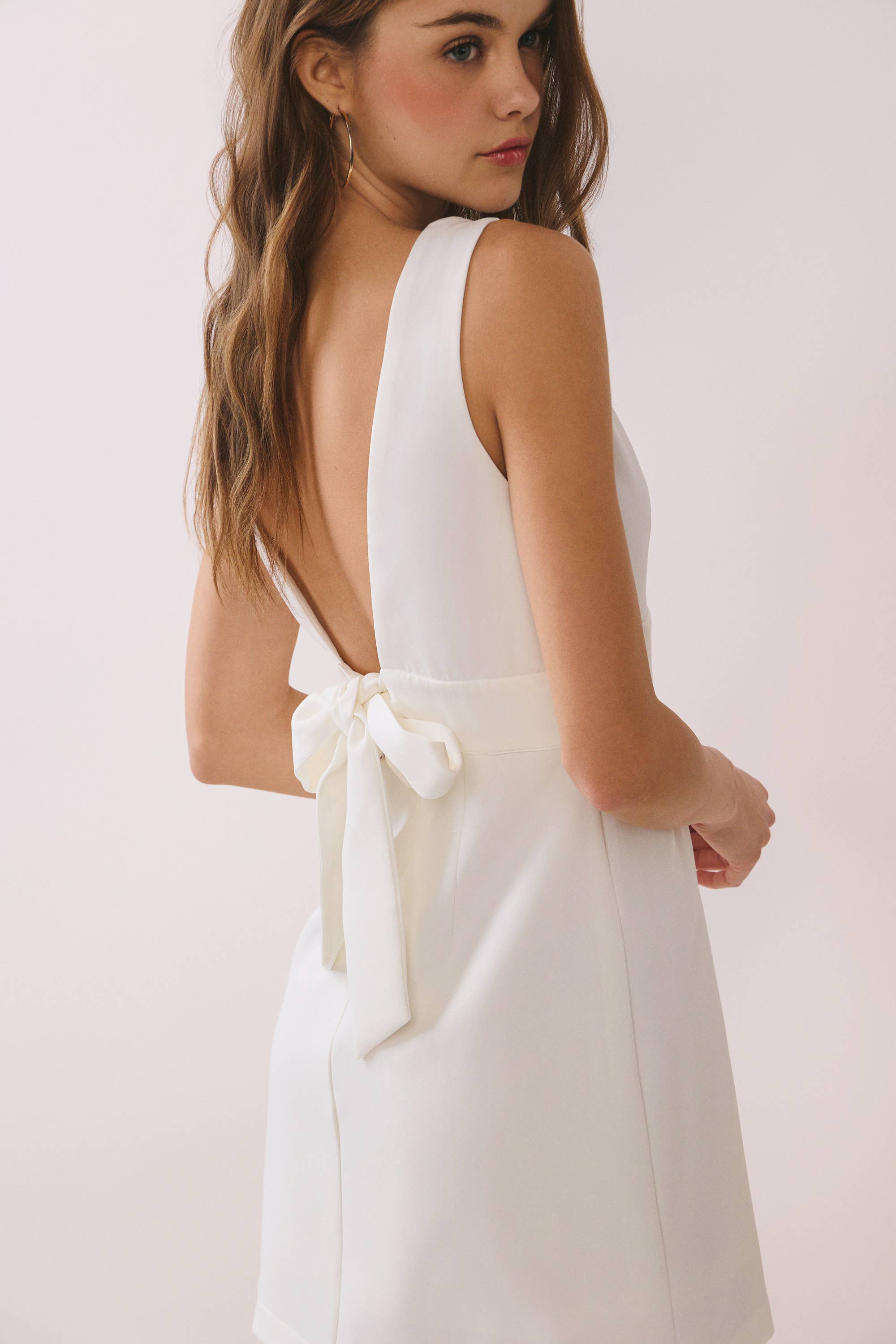 a white sleeveless mini dress with a deep V-neckline, wide waistband, dramatic low back, and self-tie bow detail