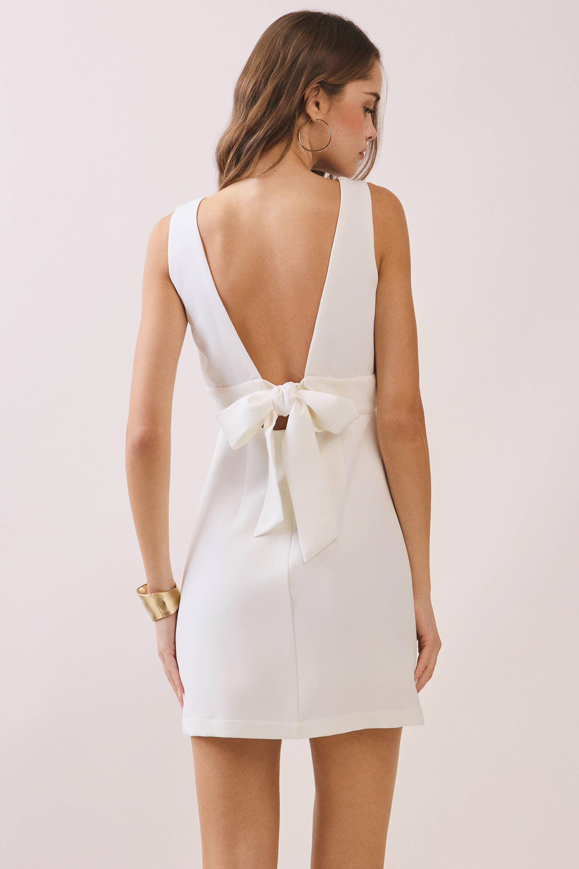 a white sleeveless mini dress with a deep V-neckline, wide waistband, dramatic low back, and self-tie bow detail