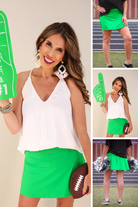 a kelly green true-to-size tennis skort designed with a slimming fit, built-in shorts, interior side pockets, smooth stretch fabric, pleated back, high rise waistband, and mini length