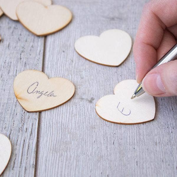 Hand writing on a heart-shaped wooden tag with other heart-shaped tags in the background.