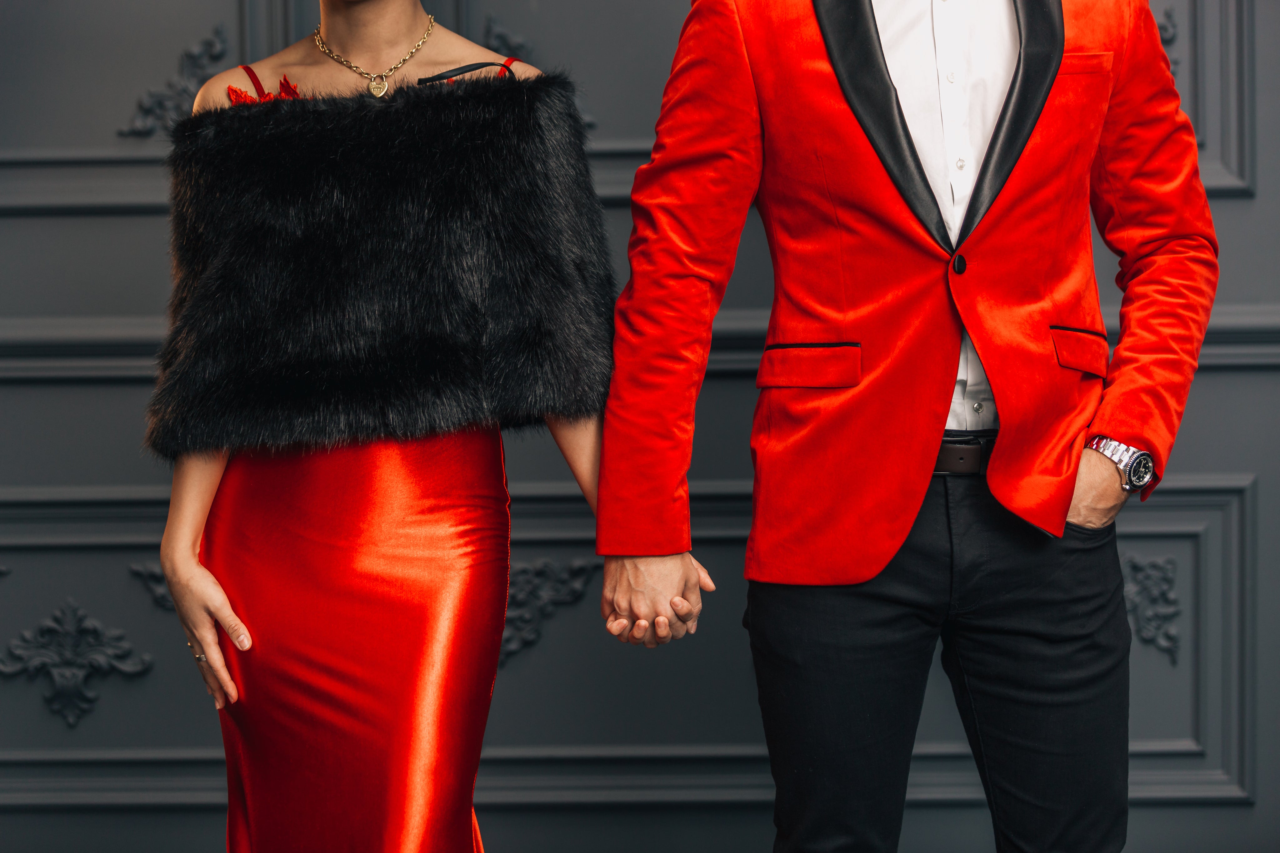 female wearing a black faux fur off the shoulder wrap over a silky red spaghetti strap dress with a man in a stunning red tux with black trim, a white dress shirt tucked into black dress pants, and a belt