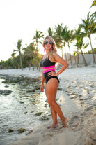 two-piece swimsuit with a scoop neck, adjustable straps, and removable padding paired with a full coverage high-waisted bottom on a solid black with a contrasting hot pink band&nbsp;