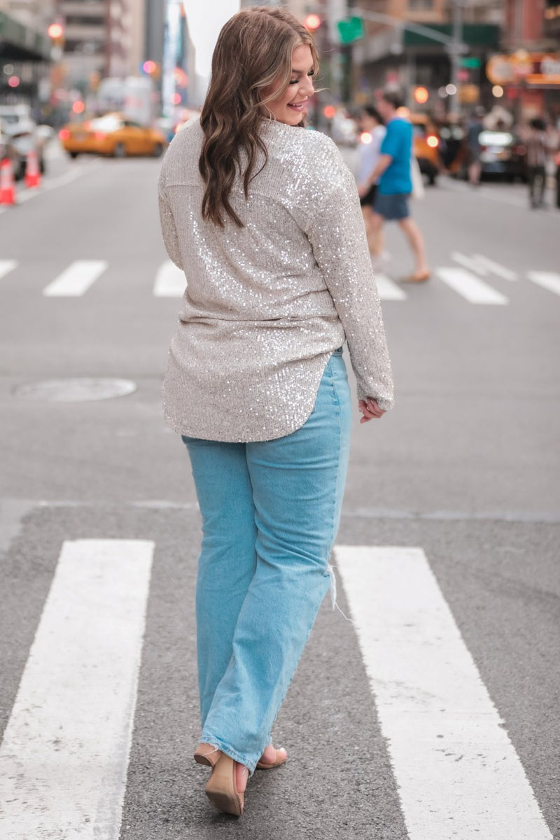 a loose long-sleeve top made of lightweight material covered in shimmering sequins with a collared neckline, fitted button closure cuffs, an accent chest pocket, and a relaxed button-up silhouette that falls into a round high-low hemline in champagne