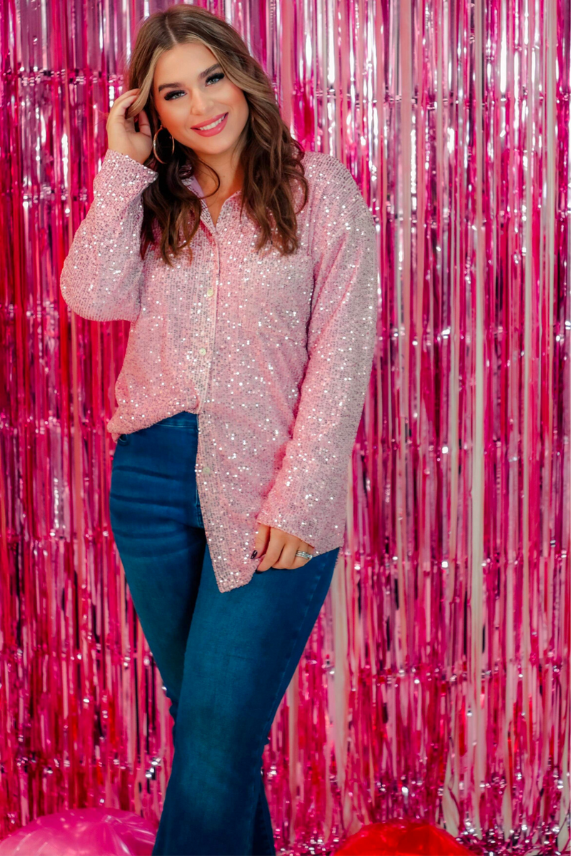 a loose long-sleeve top made of lightweight material covered in shimmering sequins with a collared neckline, fitted button closure cuffs, an accent chest pocket, and a relaxed button-up silhouette that falls into a round high-low hemline in pink